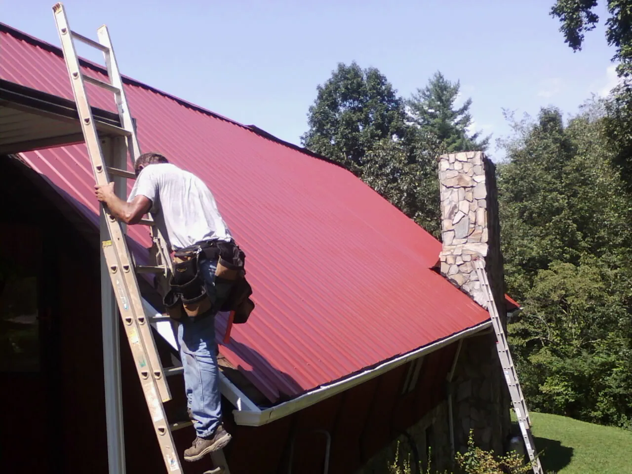 Roofer on a ladder beside a red metal roof and stone chimney — Soffit & Fascia Repair by Roofing Brothers in Compton